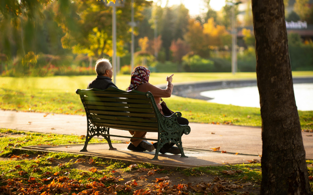 Two elderly people sit on a green ornate park bench in a sunny autumn park, one person pointing as they chat while leaves litter the ground.