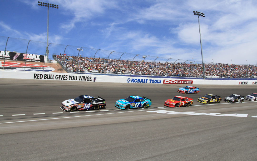 NASCAR-style race on an oval track with multiple colorful race cars speeding past spectators in the stands, under a blue sky with sponsor banners along the wall, including Red Bull and Dodge signs.